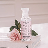 A bottle of Organic Bunny Paw Cleanser with a soft pink label featuring illustrated dog faces, placed on a white book next to a pink rose. The setting is minimal and elegant, emphasizing a natural, gentle grooming product for pets.