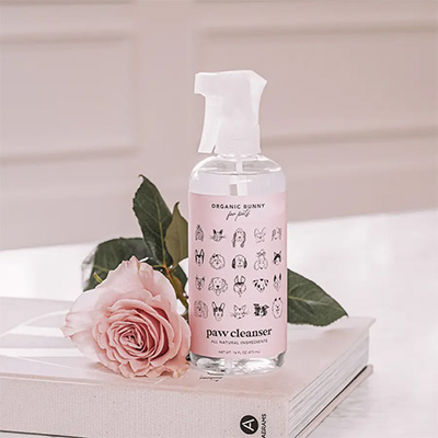 A bottle of Organic Bunny Paw Cleanser with a soft pink label featuring illustrated dog faces, placed on a white book next to a pink rose. The setting is minimal and elegant, emphasizing a natural, gentle grooming product for pets.