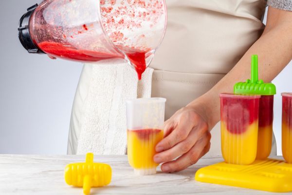 Homemade popsicles for dogs A person in a beige apron pours a red fruit mixture from a blender into a popsicle mold already partially filled with mango puree. Several finished two-tone popsicles that are safe for dogs are seen beside them in a bright yellow mold tray with green and yellow stick handles.